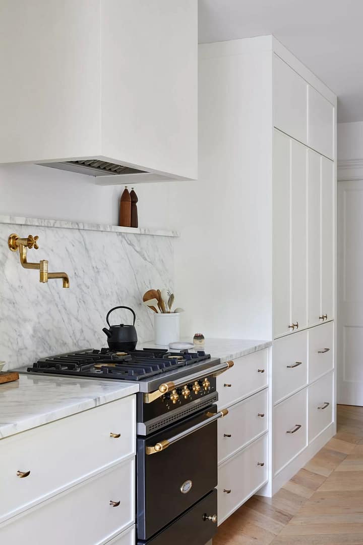 Bright modern kitchen with white cabinetry, marble backsplash, brass pot filler, and a black-gold gas range.