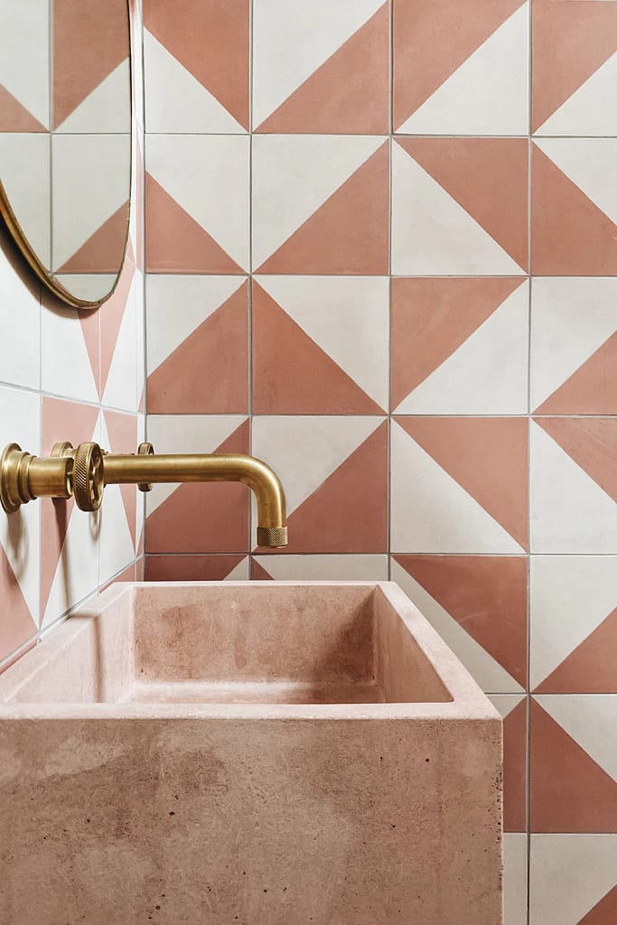 A modern bathroom renovation in North Vancouver BC by Quay Construction featuring a custom pink stone vessel sink and luxury gold wall-mounted faucets against a geometric terracotta tile backsplash.