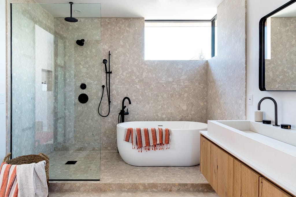 Modern bathroom from a home renovation in Hastings–Sunrise featuring a curbless glass shower, freestanding white soaking tub, and terrazzo stone walls with matte black fixtures.