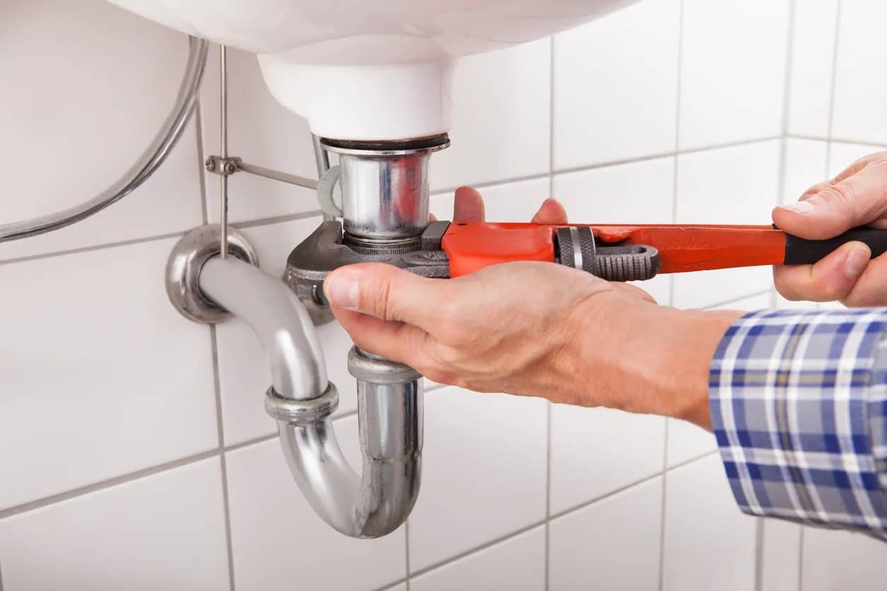 Plumber tightens sink pipes with a wrench, white square tiling behind