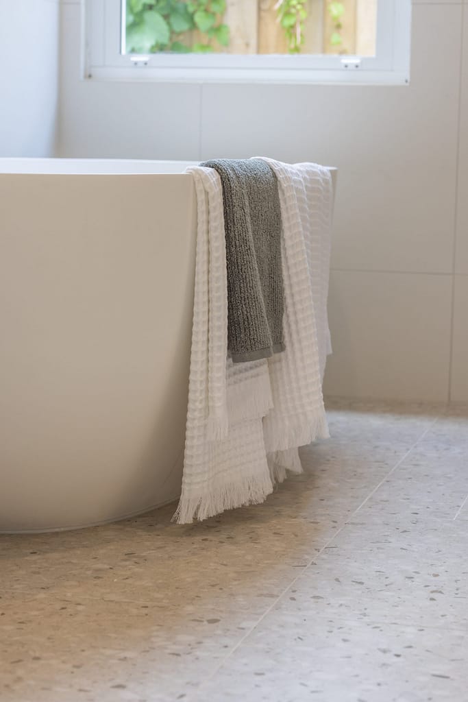 A modern bathroom renovation in West Vancouver featuring a white freestanding soaking tub with waffle-knit towels on a light grey terrazzo tile floor.
