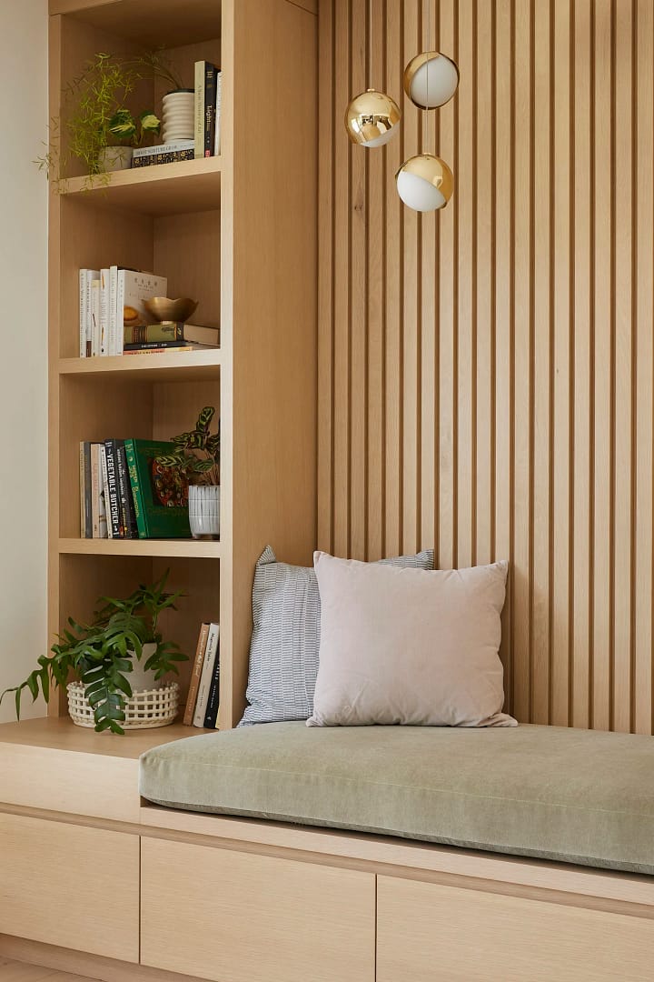 A custom-built reading nook from a home renovation in Yaletown featuring an oak slat accent wall, integrated bookshelf, and a sage green cushioned bench with gold globe pendant lights.