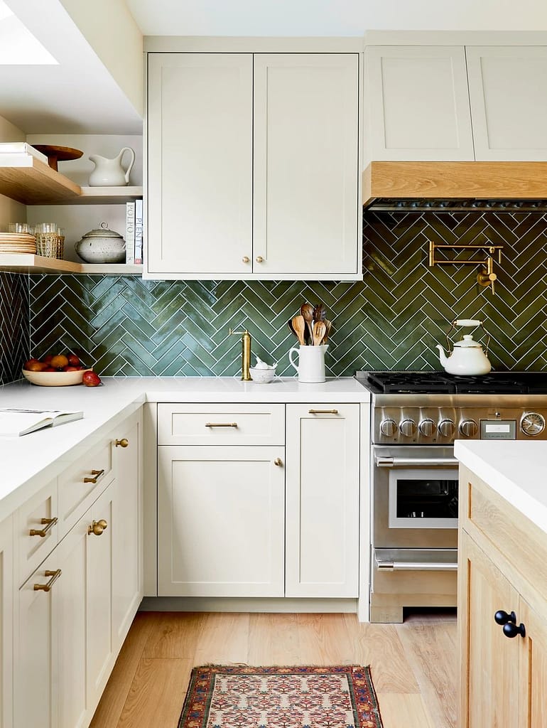 Coquitlam kitchen layout featuring dark green herringbone backsplash tiles, cream shaker cabinets, gold hardware, and open shelving in a functional corner workspace.