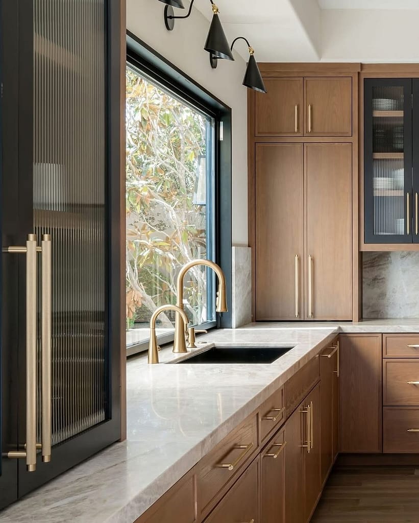 Warm modern kitchen with natural wood cabinets, gold hardware, marble countertops, and a black sink beneath a large window.