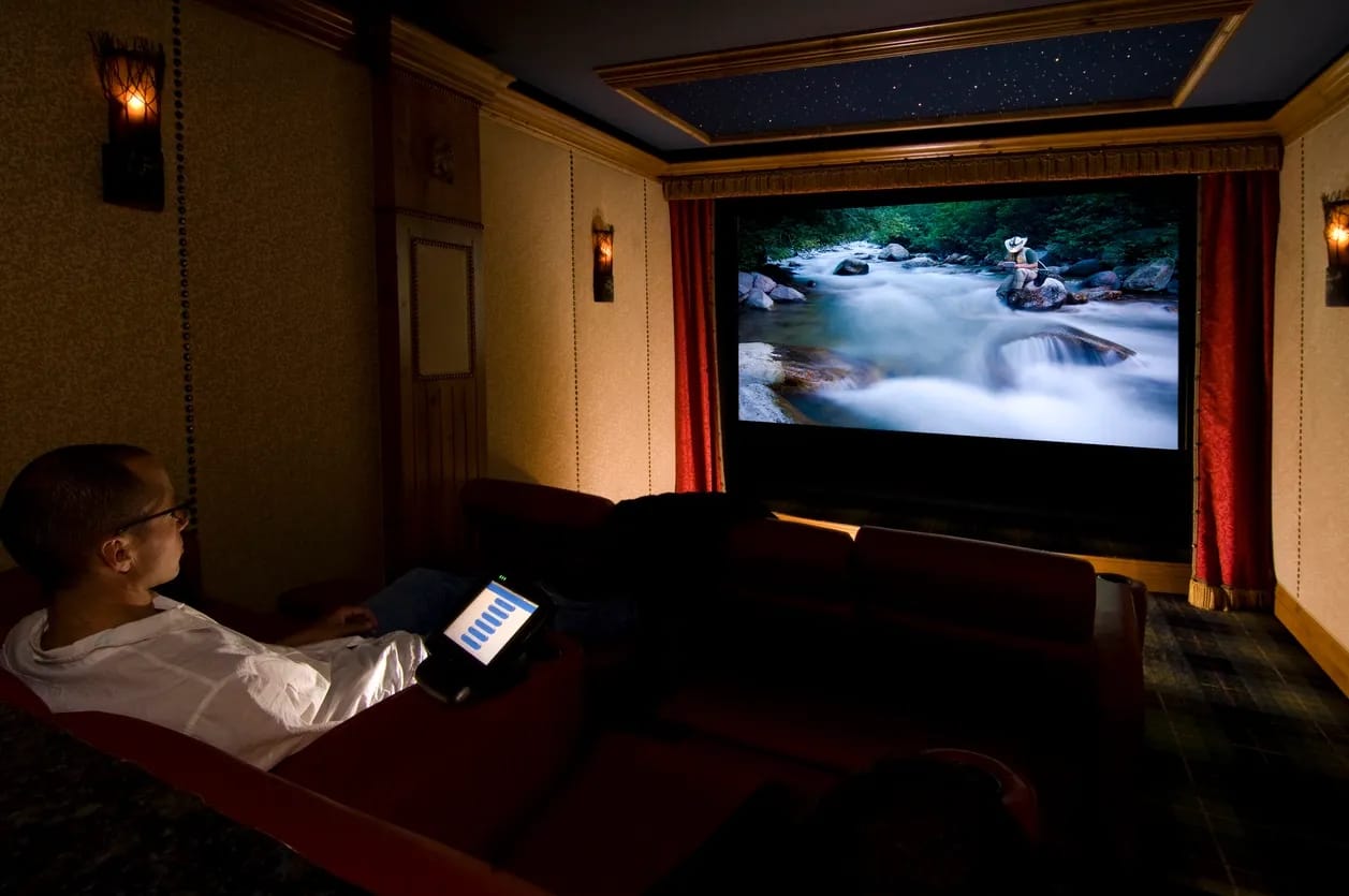 Man watches his home theatre system with red curtains around large TV and large screen remote control